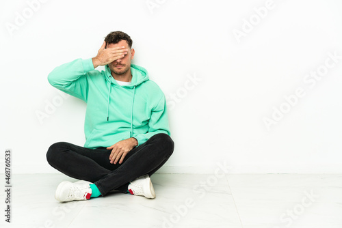 Young handsome caucasian man sitting on the floor covering eyes by hands. Do not want to see something