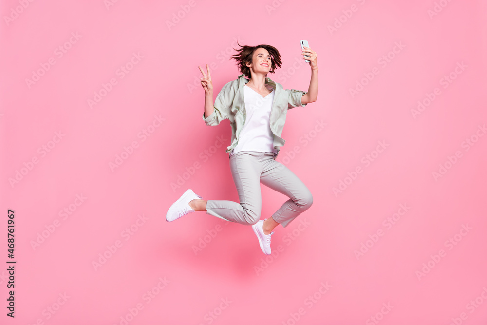 Full length photo of nice millennial lady jump with telephone v-sign wear grey green suit sneakers isolated on pink background Full length photo of nice millennial lady jump with telephone v-sign wear grey green suit sneakers isolated on pink background