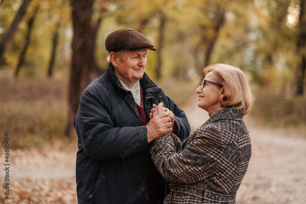 Elderly man looking at his elegant retired woman with love, warming her hands and enjoying her beauty, senior married couple dancing in autumn park, celebrating anniversary, affection Elderly man looking at his elegant retired woman with love, warming her hands and enjoying her beauty, senior married couple dancing in autumn park, celebrating anniversary, affection