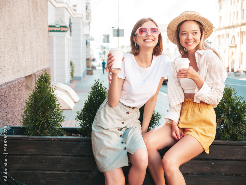 Two young beautiful smiling hipster female in trendy summer clothes.Sexy carefree women posing in the street. Positive pure models having fun at sunset. They drinking coffee or tea in plastic cup Two young beautiful smiling hipster female in trendy summer clothes.Sexy carefree women posing in the street. Positive pure models having fun at sunset. They drinking coffee or tea in plastic cup