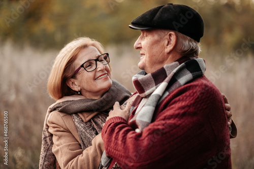 Elderly man looking at his elegant retired woman with love, warming her hands and enjoying her beauty, senior married couple dancing in autumn park, celebrating anniversary, affection