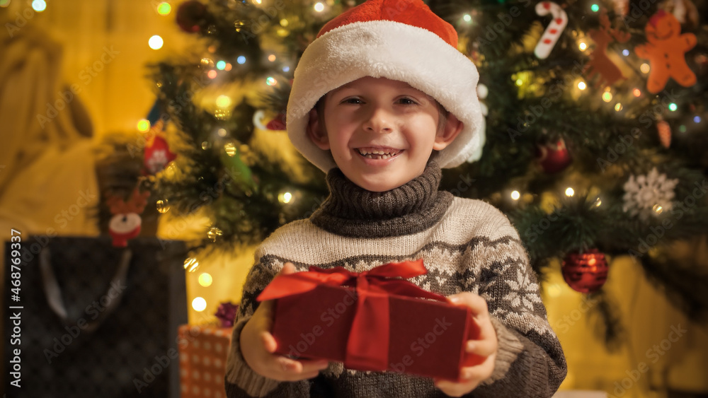 Portrait of smiling boy in Santa hat holding Christmas present box and smiling in camera. Families and children celebrating winter holidays.