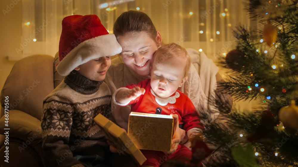 Happy family with children sitting in armchair at Christmas tree and looking inside glowing Christmas present box. Families and children celebrating winter holidays.
