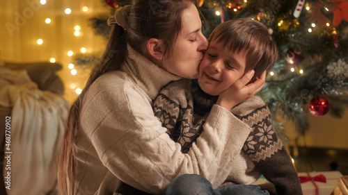 Portrait of happy mother hugging and kissing her son on Christmas eve next to glowing Christmas tree. Families and children celebrating winter holidays.