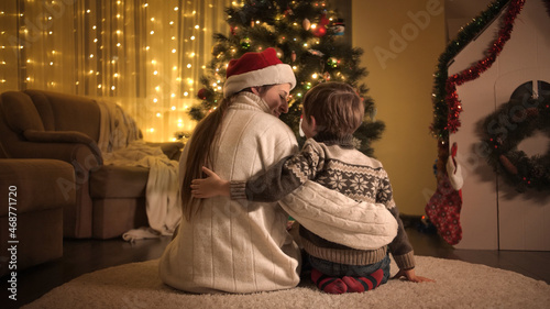 Little boy with mother sitting on floor in living room and looking on Christmas tree. Families and children celebrating winter holidays.