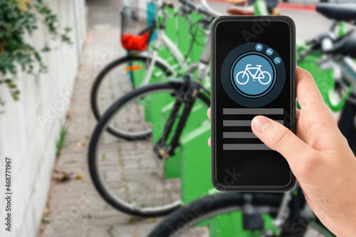 technology, transport and sustainability concept - close up of hand holding smartphone with mobile app and bicycle icon on screen over electric bike parking and charging station in city on background