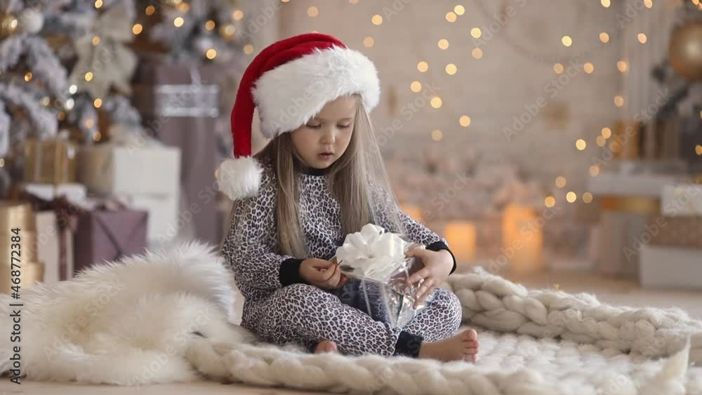Little cute girl in pajamas and santa hat at home opens her christmas gift Little cute girl in pajamas and santa hat at home opens her christmas gift