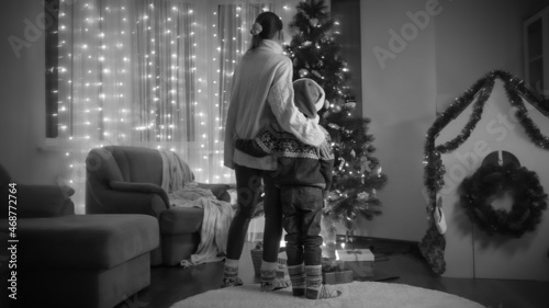 Black and white photo of mom with son in Santa hat standing next to glowing Christmas tree in living room