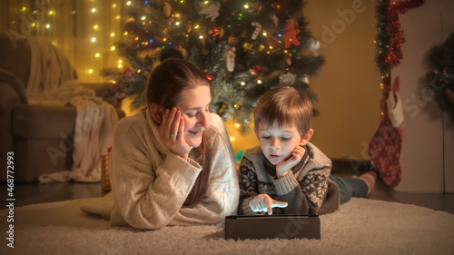 Young mother with son lying next to Christmas tree and using digital tablet. Pure emotions of families and children celebrating winter holidays.