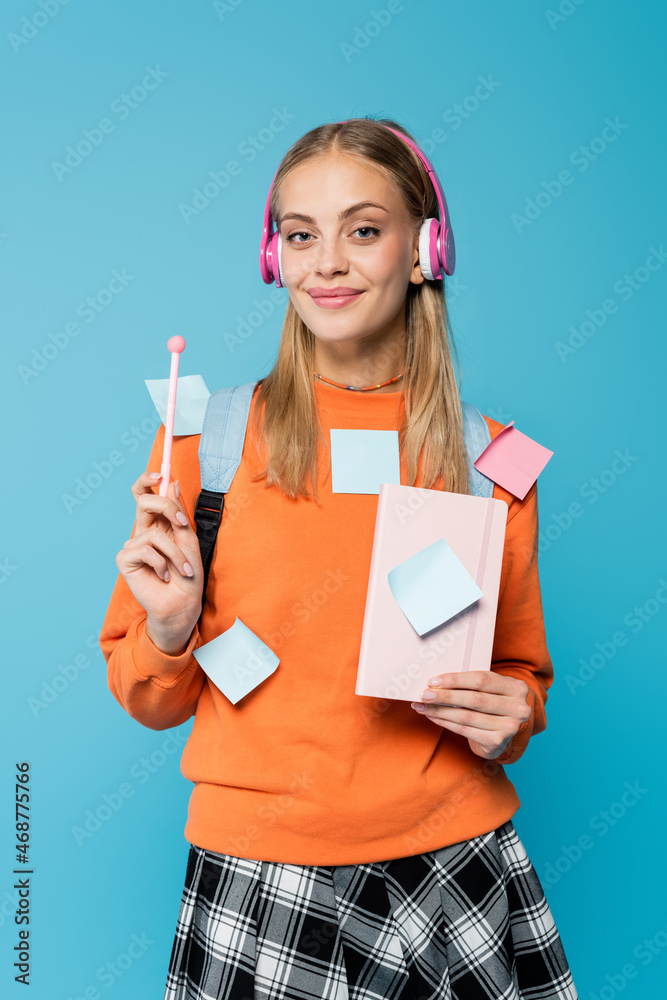 Pretty student in headphones and sticky notes holding notebook and pen isolated on blue Pretty student in headphones and sticky notes holding notebook and pen isolated on blue