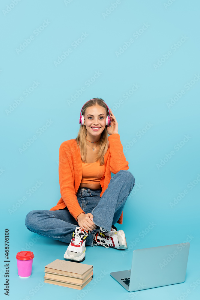 Happy blonde student in headphones sitting near laptop, paper cup and books on blue background