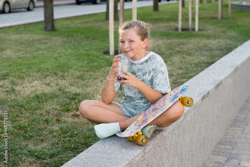 Beautiful teen girl sitting in the park with a skateboard in his hand, drinking lemonade or juice on a hot summer eternal. Leisure of teenagers without gadgets