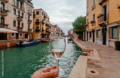 Venice with water canals and white wine glass in hand of happy traveler, Italy. Ancient italian city with old houses, bars, restaurants, narrow vessels between mansions