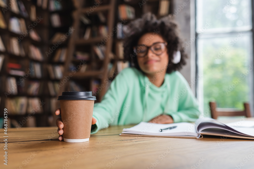 Fototapeta premium blurred african american student in eyeglasses and headphones reaching paper cup