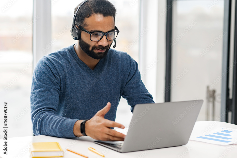 © Vadim Pastuh - Focused confident male Indian remote consultant wearing headset using laptop computer to virtual connection with customers or employees, customer service support manager talking at virtual meeting © Vadim Pastuh - Focused confident male Indian remote consultant wearing headset using laptop computer to virtual connection with customers or employees, customer service support manager talking at virtual meeting