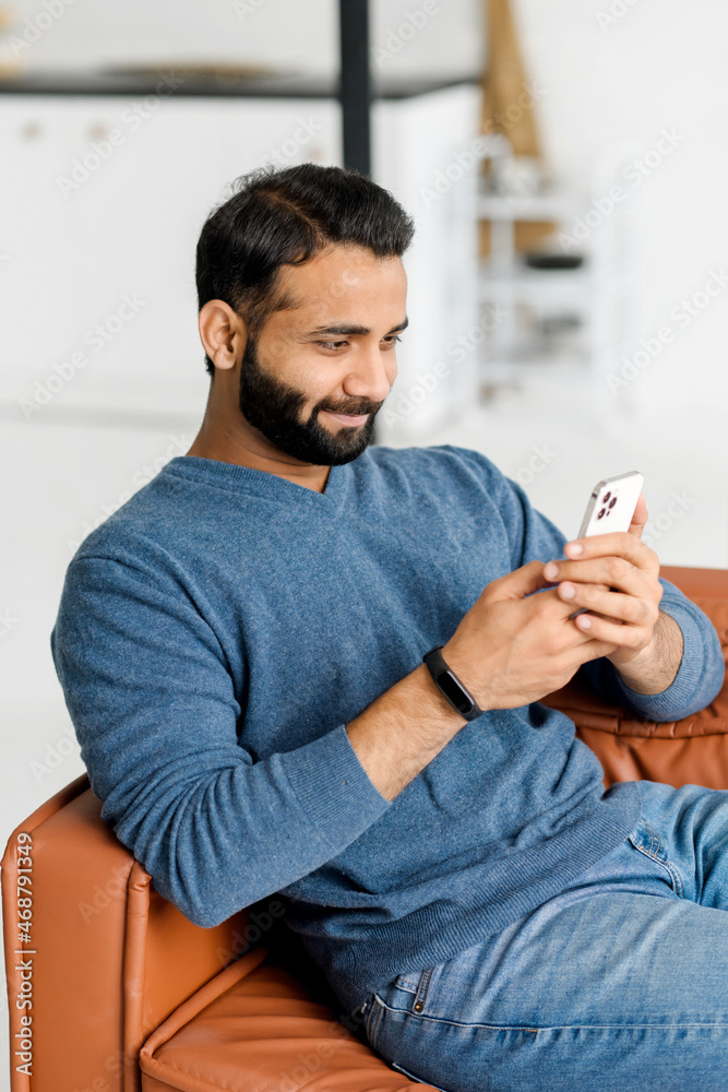 Serene Indian man holds smartphone sitting on the couch at home, chatting online with friends, spend leisure time in social media, texting, multiracial guy scrolling news feed, shopping online