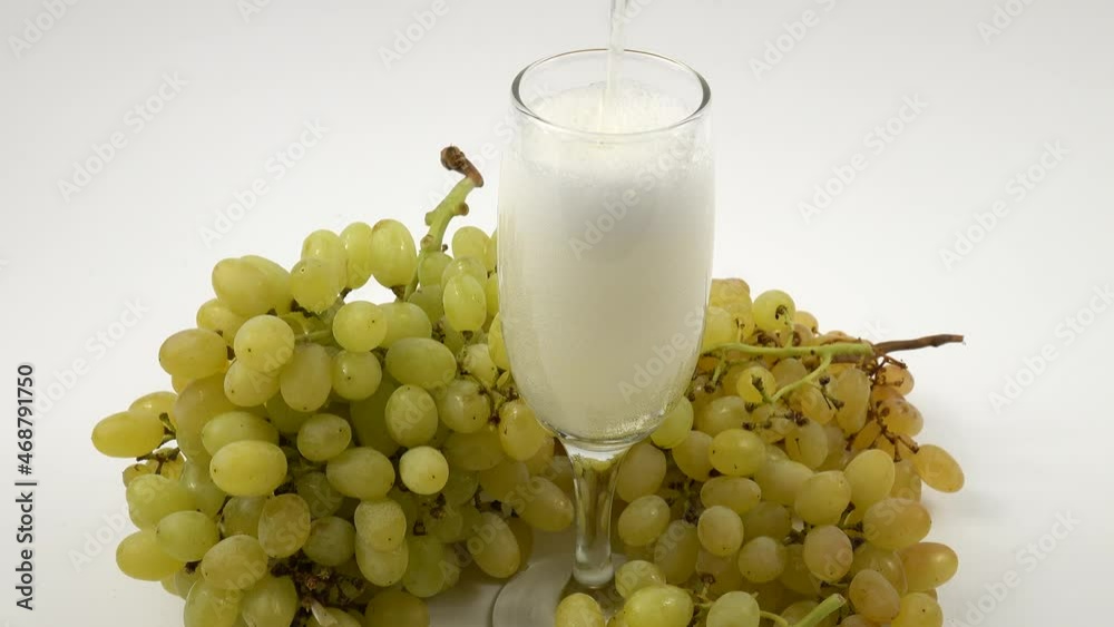 White champagne is poured into a transparent glass, standing on a white table near a large bunch of ripe white grapes, background White champagne is poured into a transparent glass, standing on a white table near a large bunch of ripe white grapes, background
