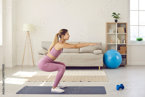 Fit young woman in sportswear do squats train exercise at home. Millennial girl workout do sports in living room, follow healthy lifestyle. Female coach or trainer working out on lockdown indoors. photo