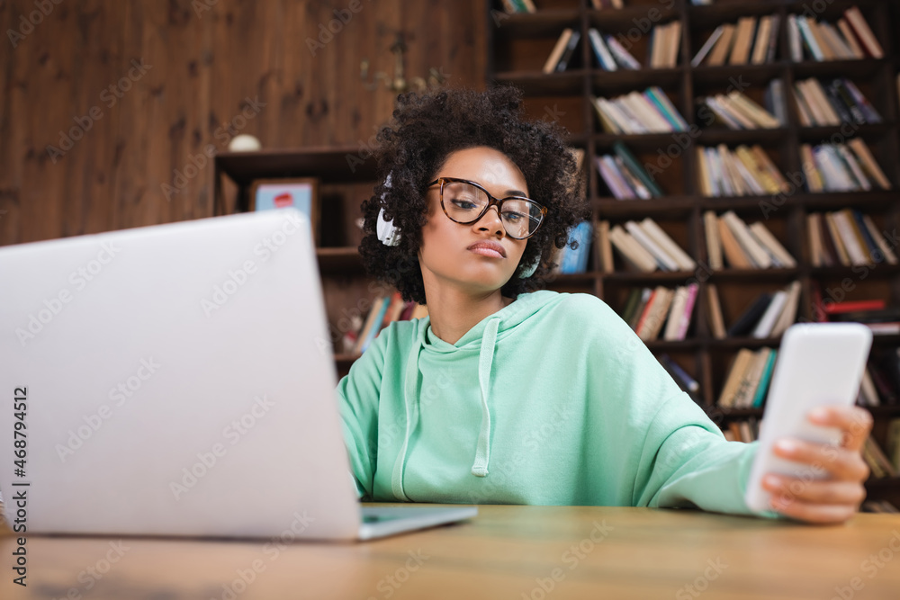 african american student in eyeglasses using smartphone near blurred laptop african american student in eyeglasses using smartphone near blurred laptop