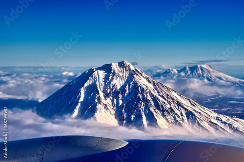 View of the snow-capped volcano from the aircraft s illuminator. Peak dormant volcano above the clouds.