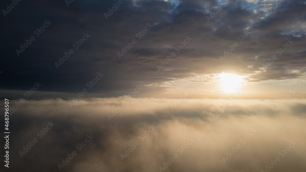 Aerial View. Flying in fog, fly in mist over the early morning clouds in the rising sun. Aerial camera shot. Flight above the clouds towards the sun with the mist clouds floating by. Misty weather