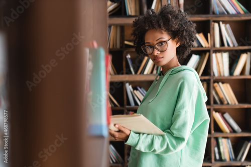 young african american woman in eyeglasses looking at camera while holding book near bookshelf