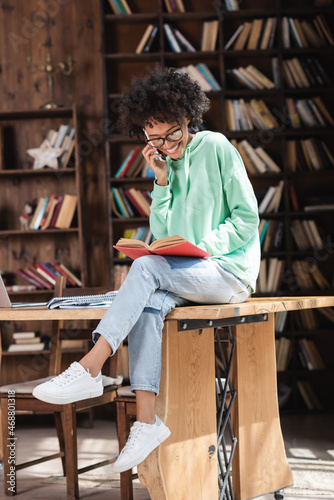 happy african american student in eyeglasses talking on cellphone and reading book while sitting on desk