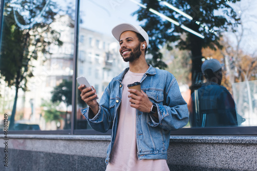 Happy Spanish hipster guy in bluetooth headphones enjoying positive music playlist from mobile radio using 4g wireless in city, cheerful male tourist in earbuds holding coffee to go and smartphone