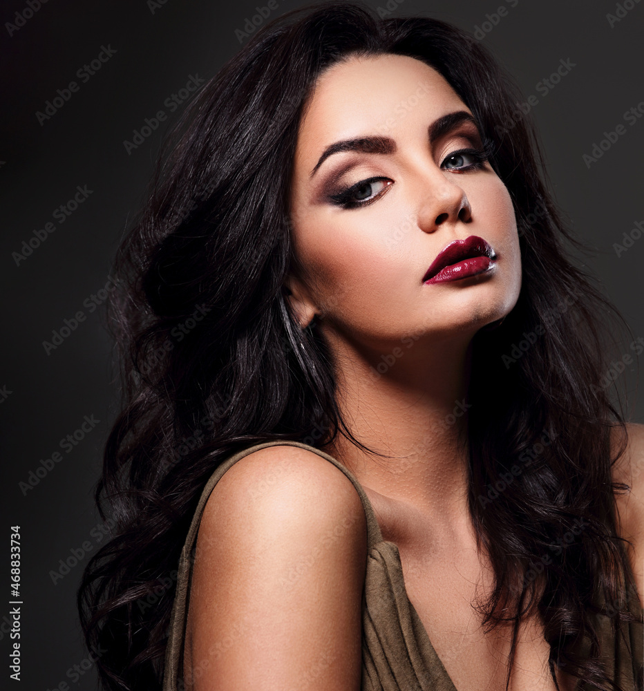 Fototapeta premium Beautiful bright makeup woman with long black curly hair style, burgundy lipstick with vamp look on dark grey background. Closeup