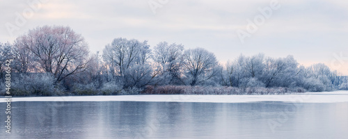 Billede på lærred Frost-covered trees on the banks of an ice-bound river in the morning during sun