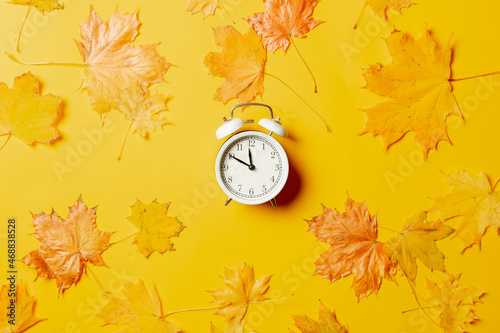 White alarm clock with maple leaves around on yellow background.