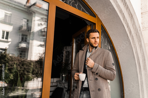 Handsome man with cup of coffee posing in coat in cafe. Male lifestyle.
