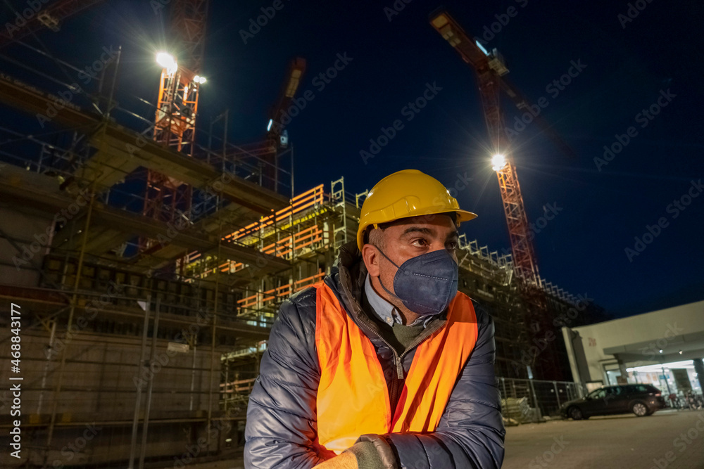 Engineering during COVID-19 Coronavirus outbreak. Occupational safety and health and protection. Construction worker posing at night wearing safety vest, hardhat and medical mask