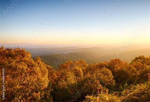 View at sunrise from a East side vista on Talimena Scenic Byway in Ouachita National Forest. Bright fall foliage in foreground trees, and heavy fog and haze between hilltops contrasting the layers.