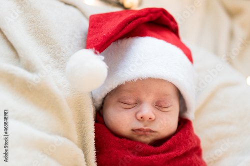 Top View Portrait First Days Of Life Newborn Cute Funny Sleeping Child Baby In Santa Hat Wrapped In Red Diaper At White Garland Background. Merry Christmas, Happy New Year, Infant, Childhood, Winter