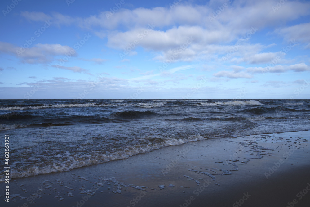 Seaside of the Baltic Sea. Contrast of bright and cloudy blue sky and a dark blue wavy sea foaming on the beach