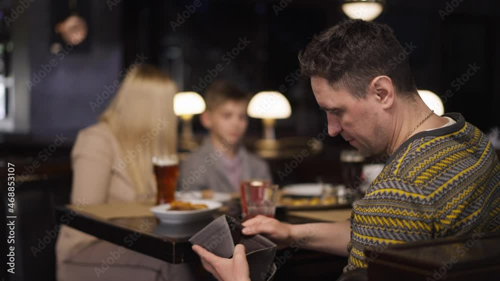 Dissatisfied man looking in empty wallet dining with family in restaurant shaking head. Portrait of stressed Caucasian father husband with wife and son indoors spending money. Slow motion Dissatisfied man looking in empty wallet dining with family in restaurant shaking head. Portrait of stressed Caucasian father husband with wife and son indoors spending money. Slow motion