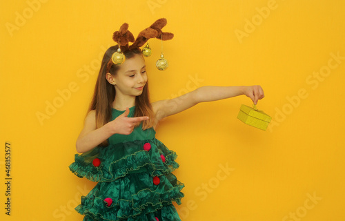 Girl with a deer ears in green festive dress holding a gift on yellow background with copy space. Christmas concept.
