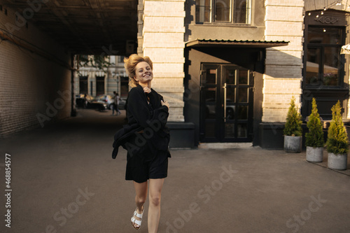 Happy caucasian young girl runs down city street smiling broadly at camera. Her short blonde hair blows in wind, she is dressed in black suit with shorts and white open shoes.