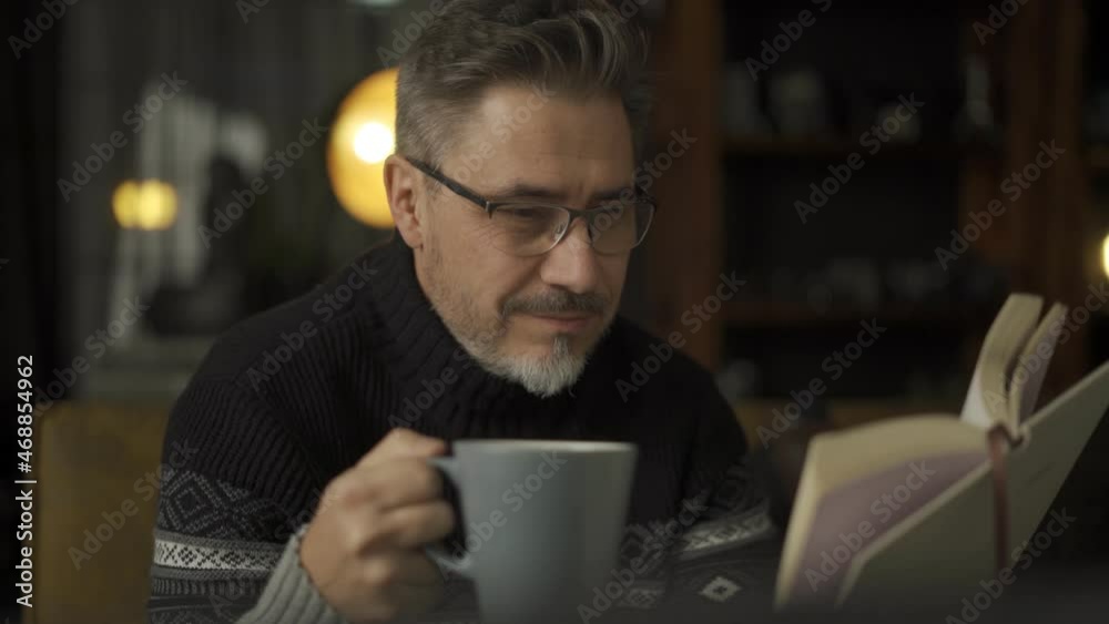 Older man sitting on sofa in dark room drinking hot tea or coffee and reading book Mature age, middle age, mid adult casual man in 50s, confident happy smiling. Cosy winter evening.