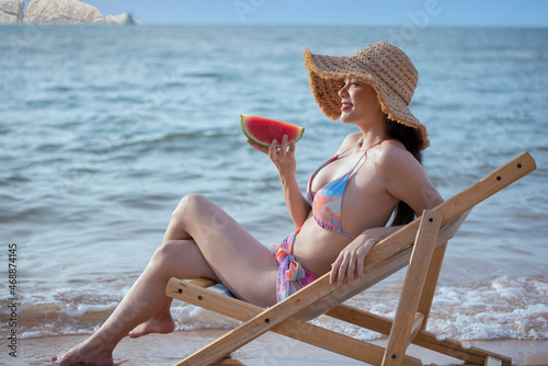Side view of beautiful asian woman  in bikini holding a watermelon sitting on a beach chair happily on summer vacation