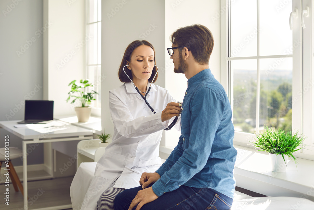 Young man visiting clinic to check heart or lungs health. Serious female doctor sitting on examination couch in modern exam room, holding stethoscope, listening to male patient's breath or heartbeat - obrazy, fototapety, plakaty 