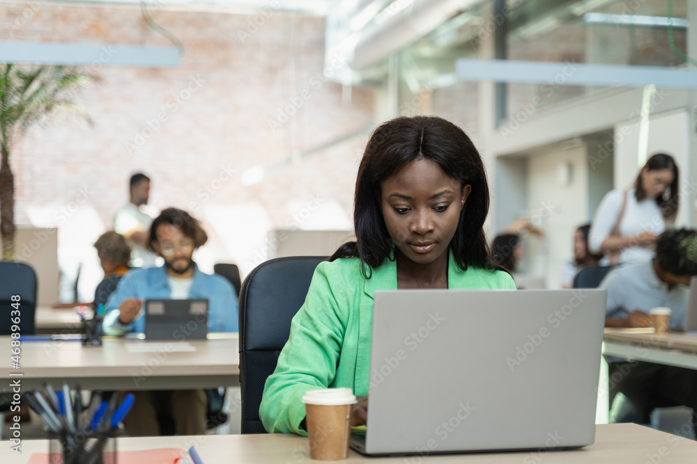 Focused young African businesswoman working online at her desk using a laptop in a bright modern office with colleagues in the background Focused young African businesswoman working online at her desk using a laptop in a bright modern office with colleagues in the background
