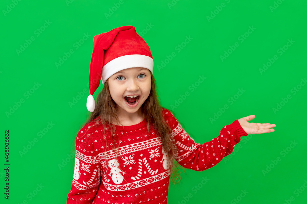 a happy little girl child in winter clothes, a Santa Claus hat and a red sweater on a green isolated background shows her fingers up at something and smiles, a place or space for text a happy little girl child in winter clothes, a Santa Claus hat and a red sweater on a green isolated background shows her fingers up at something and smiles, a place or space for text