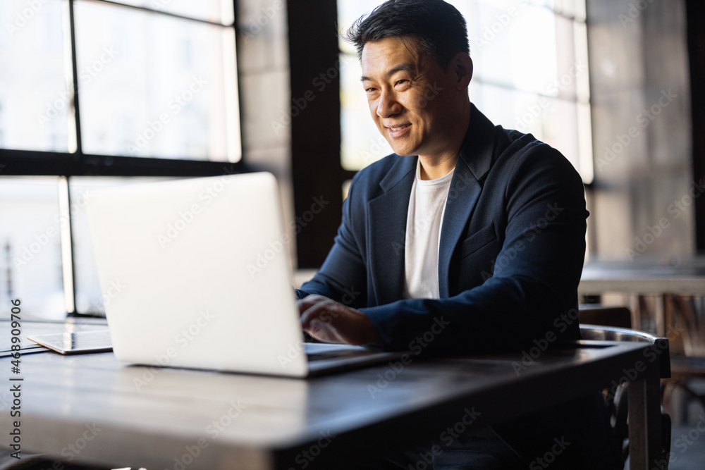 Asian businessman typing on laptop during work in cafe. Concept of remote and freelance work. Smiling adult successful man wearing suit and glasses sitting at wooden desk. Sunny day Asian businessman typing on laptop during work in cafe. Concept of remote and freelance work. Smiling adult successful man wearing suit and glasses sitting at wooden desk. Sunny day