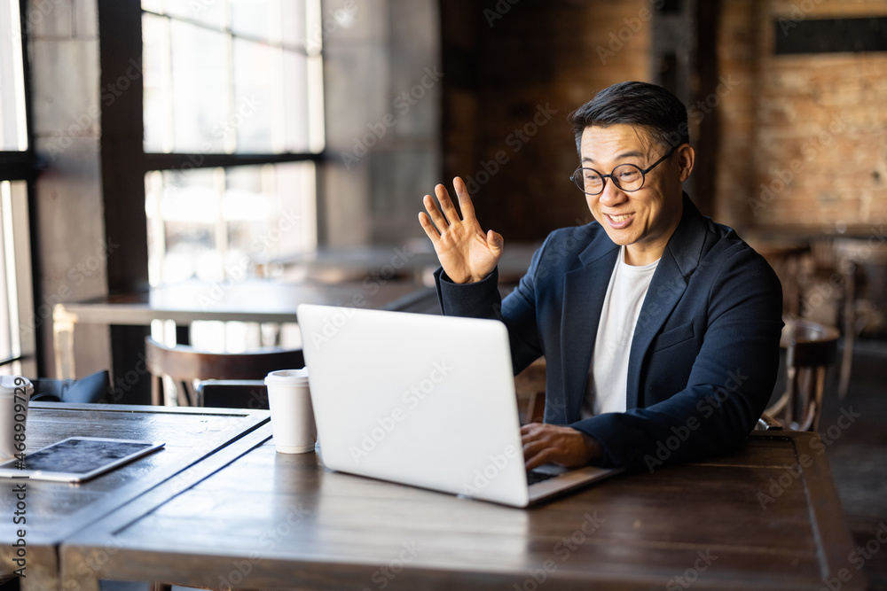 Asian businessman having a video call on laptop in cafe. Concept of remote and freelance work. Smiling adult successful man wearing suit and glasses sitting at wooden desk. Sunny day Asian businessman having a video call on laptop in cafe. Concept of remote and freelance work. Smiling adult successful man wearing suit and glasses sitting at wooden desk. Sunny day