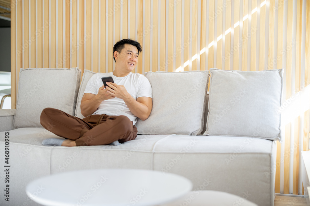 Asian man sitting on sofa, using smartphone and looking in sunny window. Concept of domestic lifestyle. Smiling adult male person wearing casual clothes. Interior of living room in modern apartment Asian man sitting on sofa, using smartphone and looking in sunny window. Concept of domestic lifestyle. Smiling adult male person wearing casual clothes. Interior of living room in modern apartment