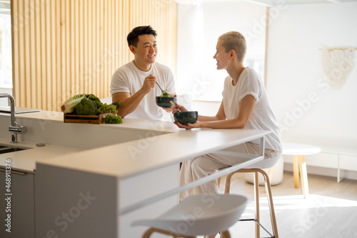 Smiling multiracial couple eating salad from fresh vegetables at home. Concept of healthy and vegetarian eating. Idea of domestic lifestyle. Asian man and european girl looking at each other