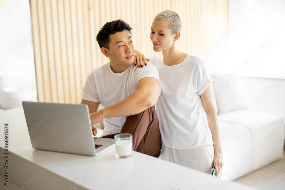 Sly caucasian woman watching something on laptop of her asian boyfriend. Concept of relationship and spending time together. Idea of domestic lifestyle. Multiracial couple in modern apartment Sly caucasian woman watching something on laptop of her asian boyfriend. Concept of relationship and spending time together. Idea of domestic lifestyle. Multiracial couple in modern apartment
