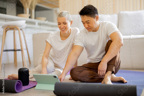 Multiracial couple sit on fitness mats and watch something on digital tablet at home. Asian man and caucasian girl reading to practising yoga. Concept of healthy lifestyle. Domestic hobby and leisure